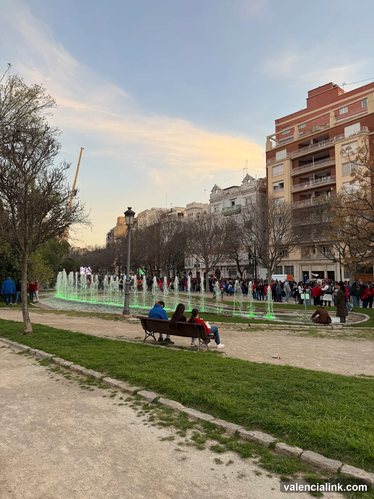 People sitting on a bench near a fountain in a park with buildings in the background.