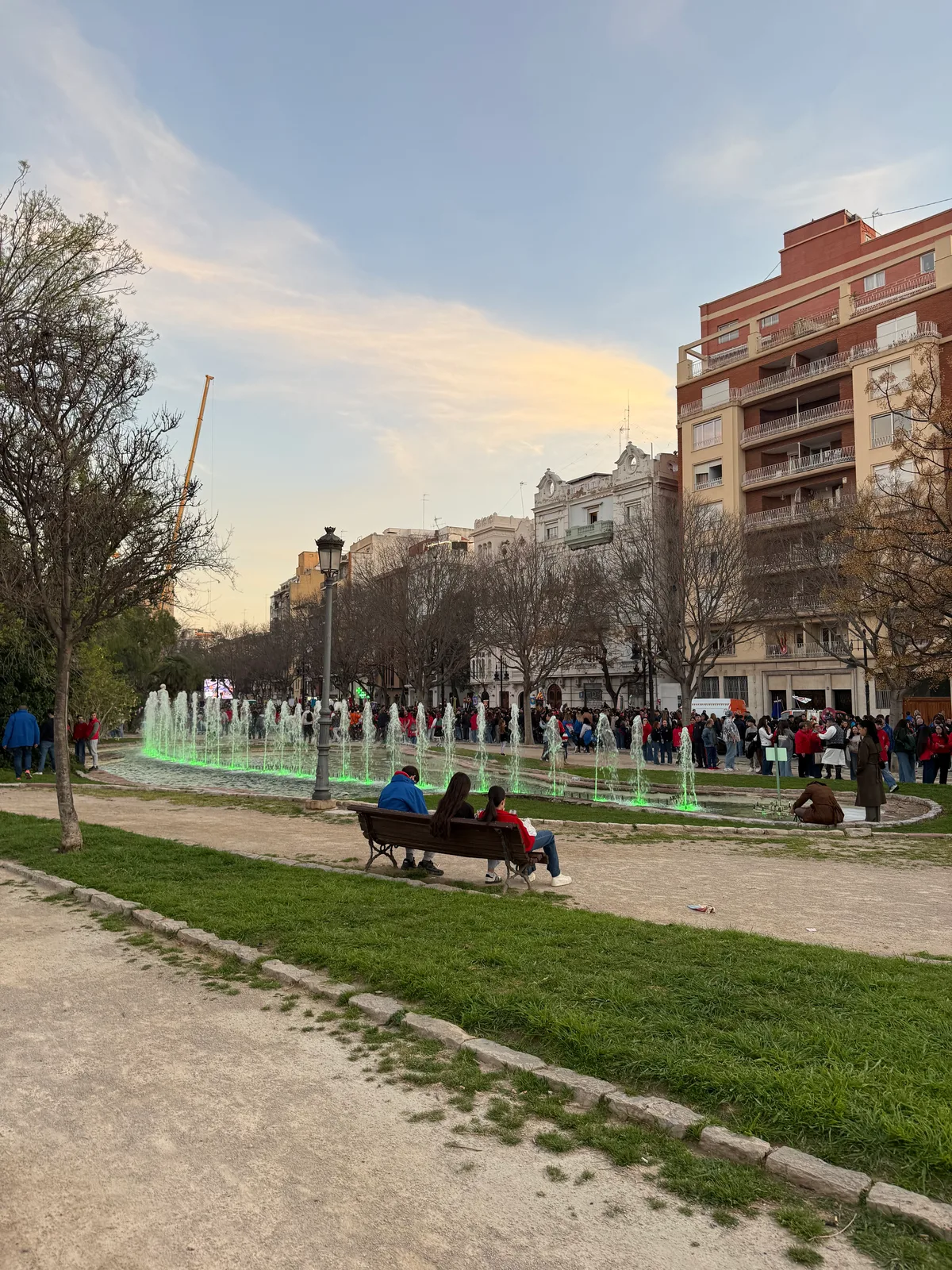 People sitting on a bench near a fountain in a park with buildings in the background.