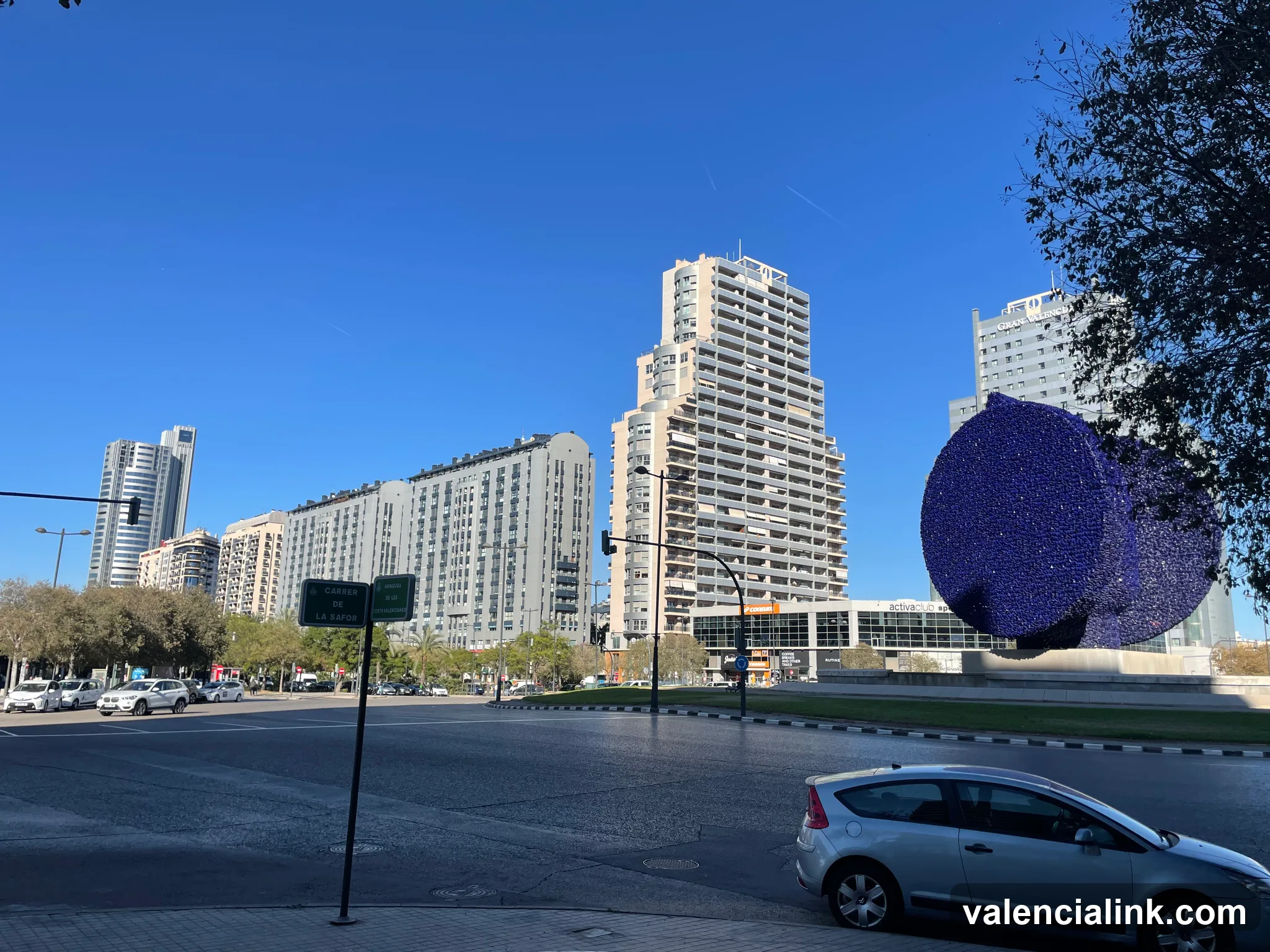 A modern urban scene with a large blue circular sculpture, tall buildings, and a clear blue sky.