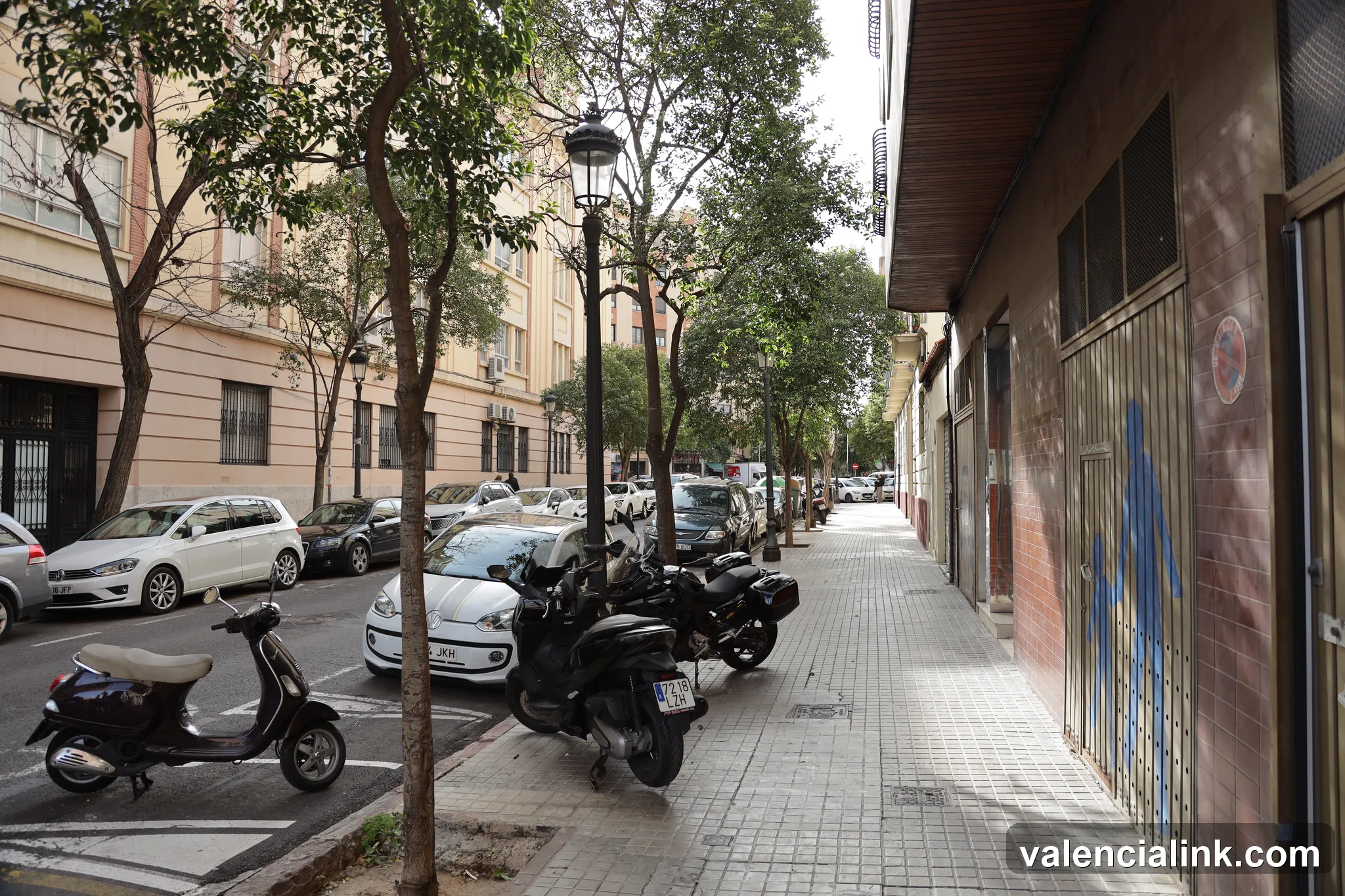 A street scene with parked scooters and cars, trees, and buildings.