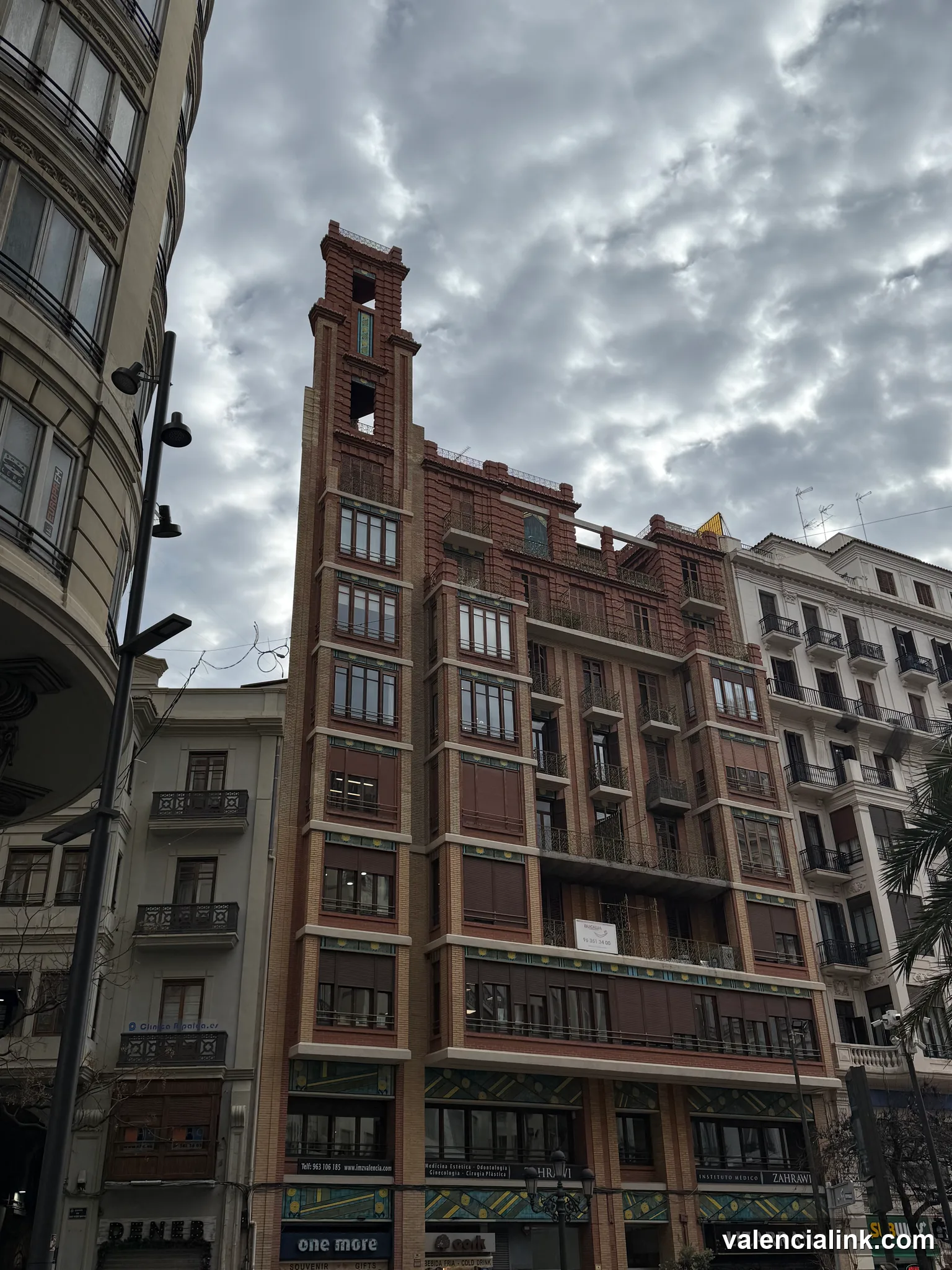 A red brick building with a tall tower and multiple balconies under a cloudy sky.