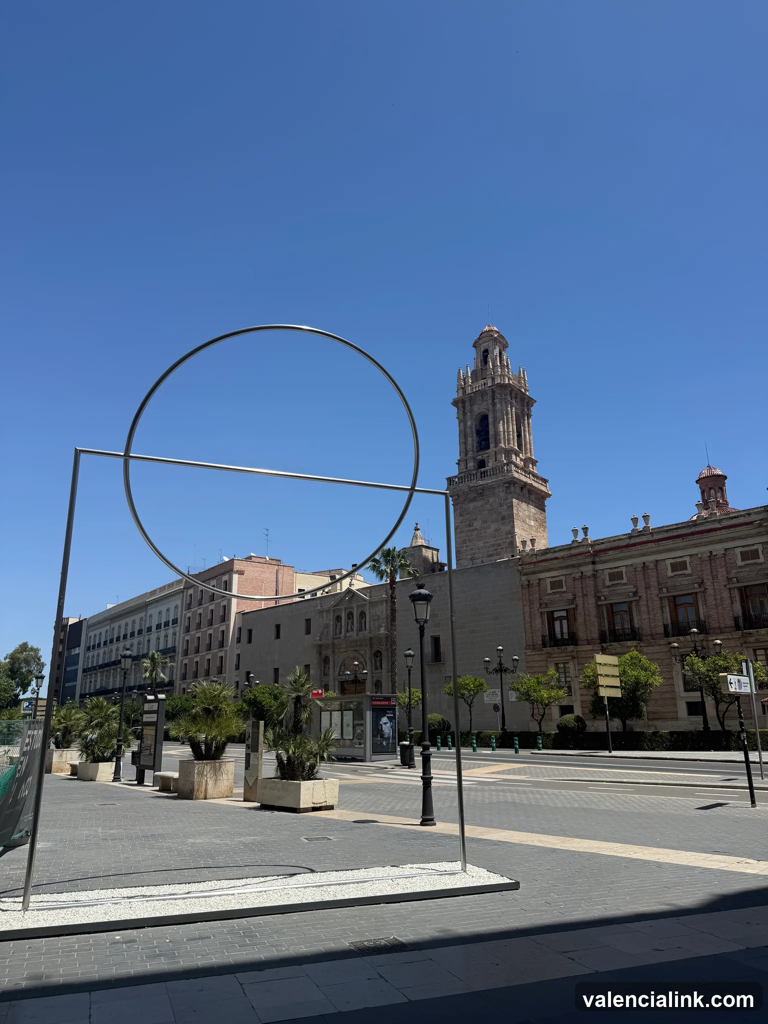 Modern art installation with a large circular sculpture in a city square with historic buildings and a clear blue sky.