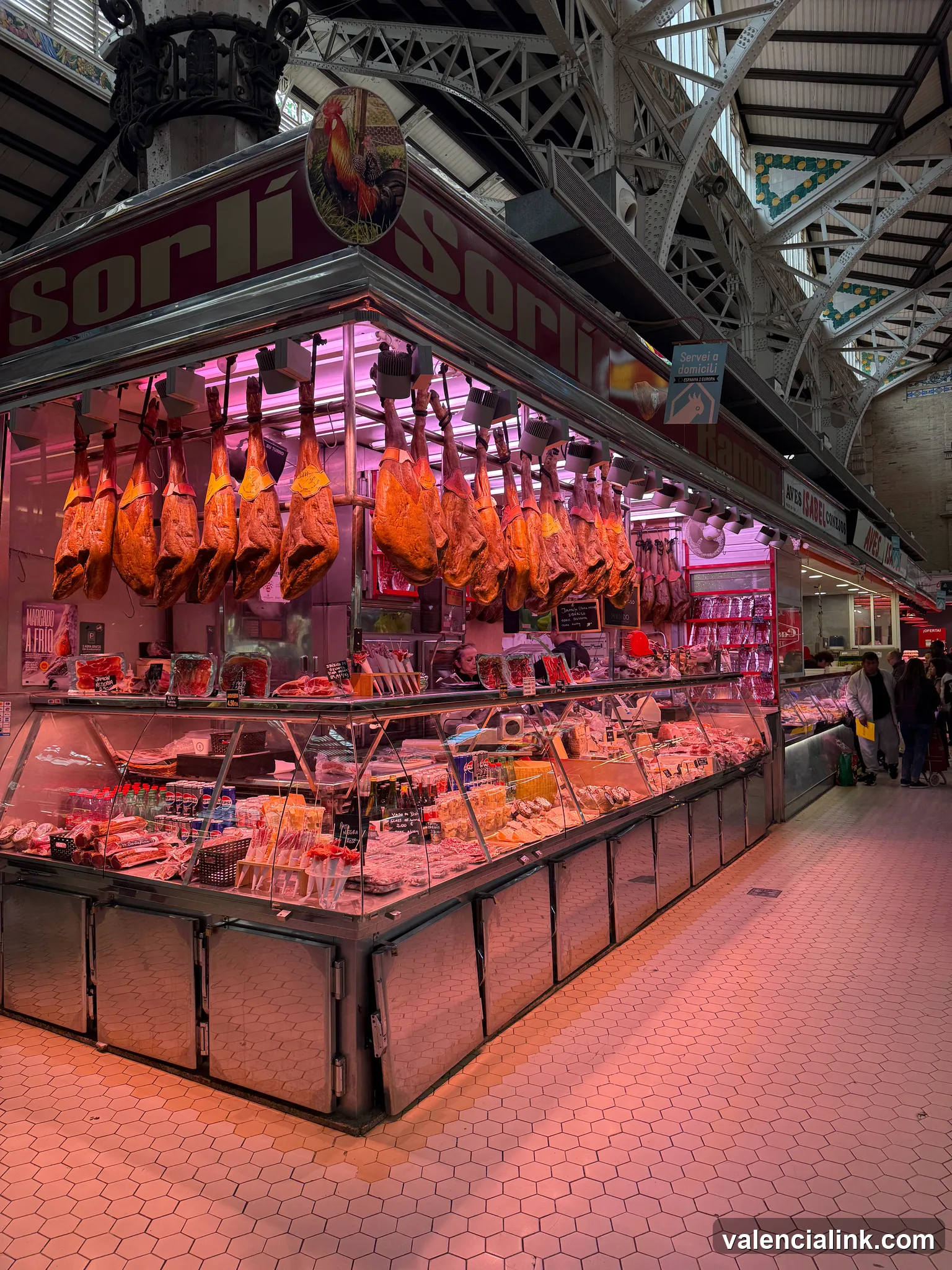 Market stall with hanging cured meats and food items under a metal archway.