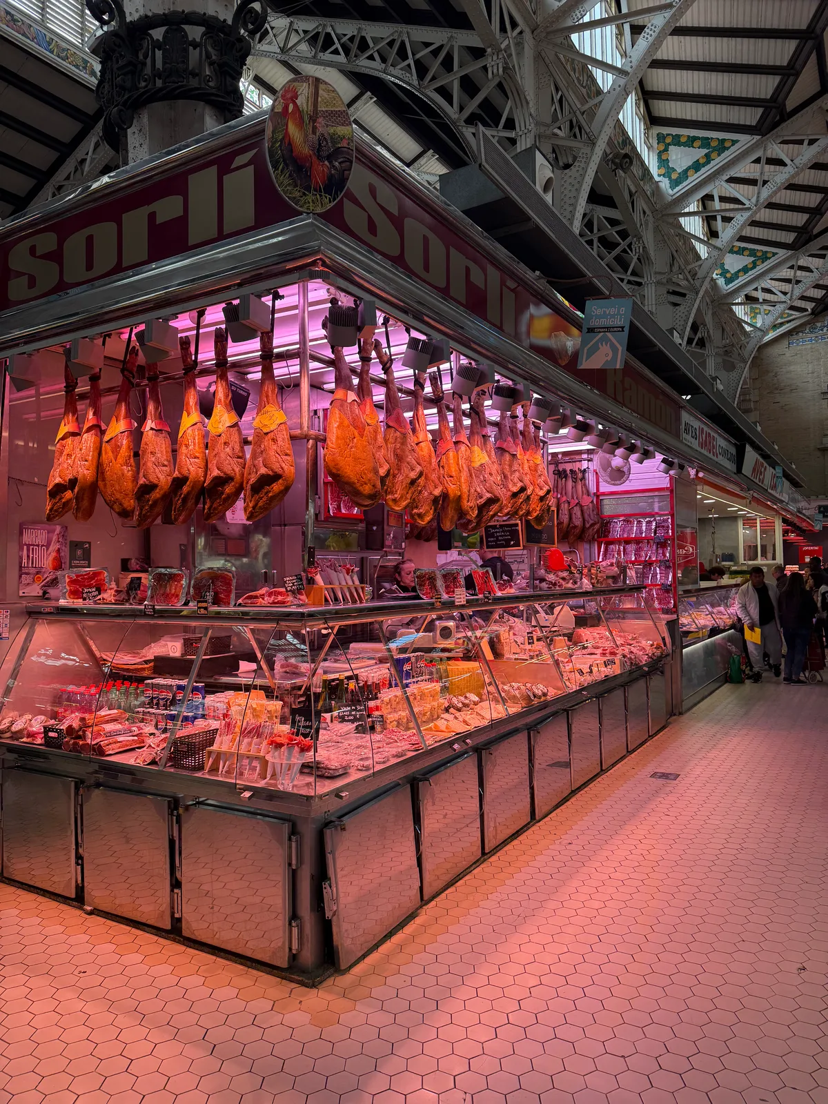 Market stall with hanging cured meats and food items under a metal archway.