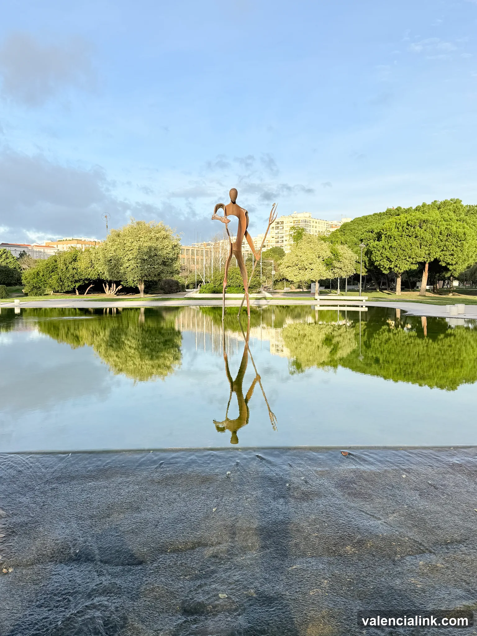 A sculpture of a figure on stilts reflecting in a pond with greenery and buildings in the background.