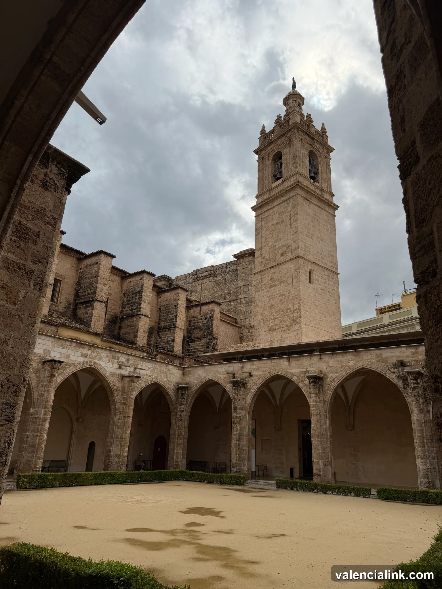 Cortile in pietra con passaggi ad arco e alto campanile sotto un cielo nuvoloso.