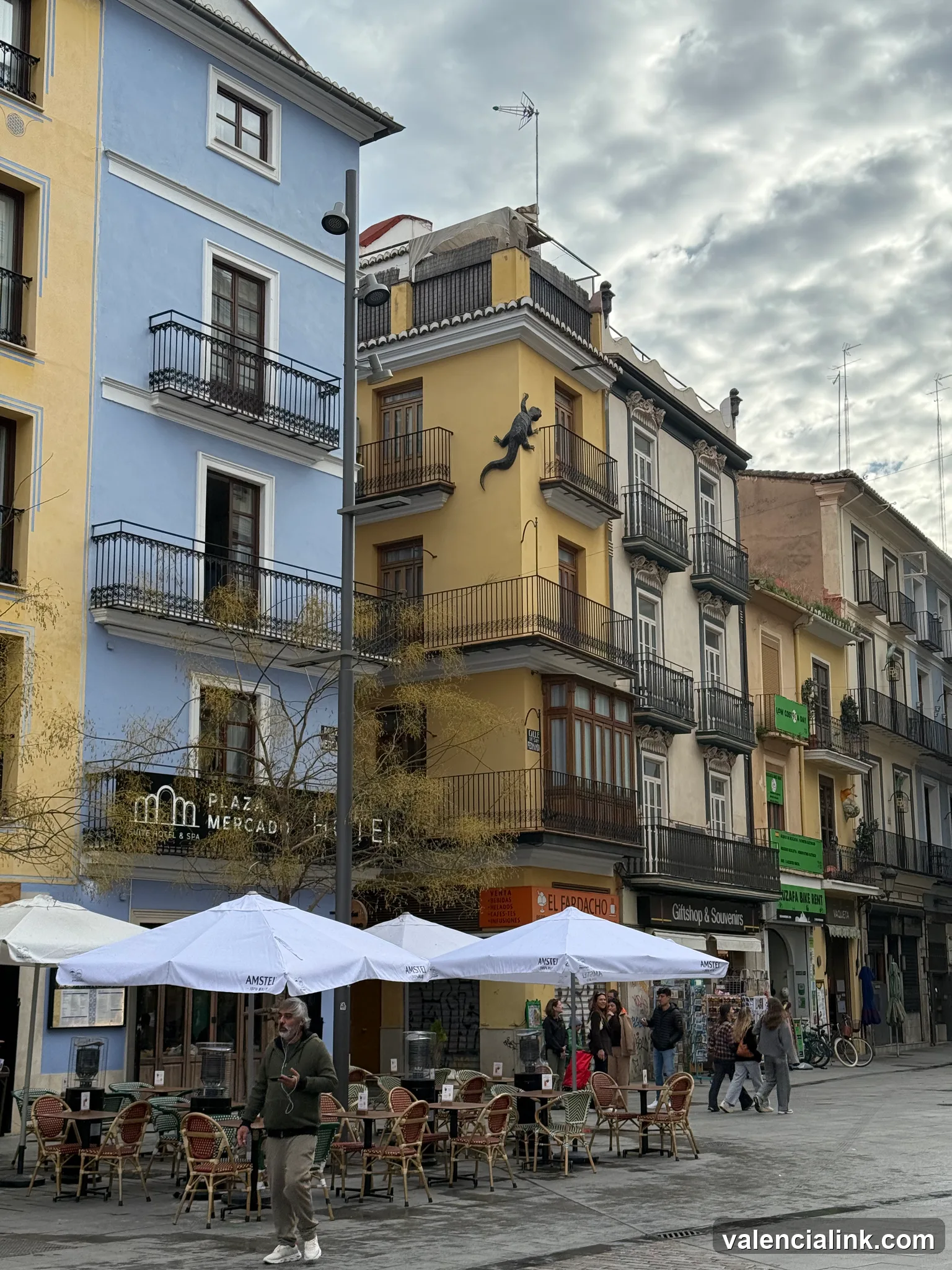 Street scene with colorful buildings, outdoor cafes, and pedestrians under a cloudy sky.