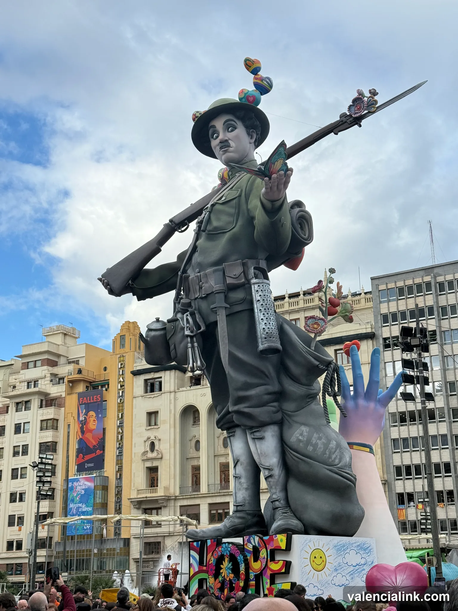 A large statue of a soldier holding a rifle in a public square with buildings and a crowd in the background.