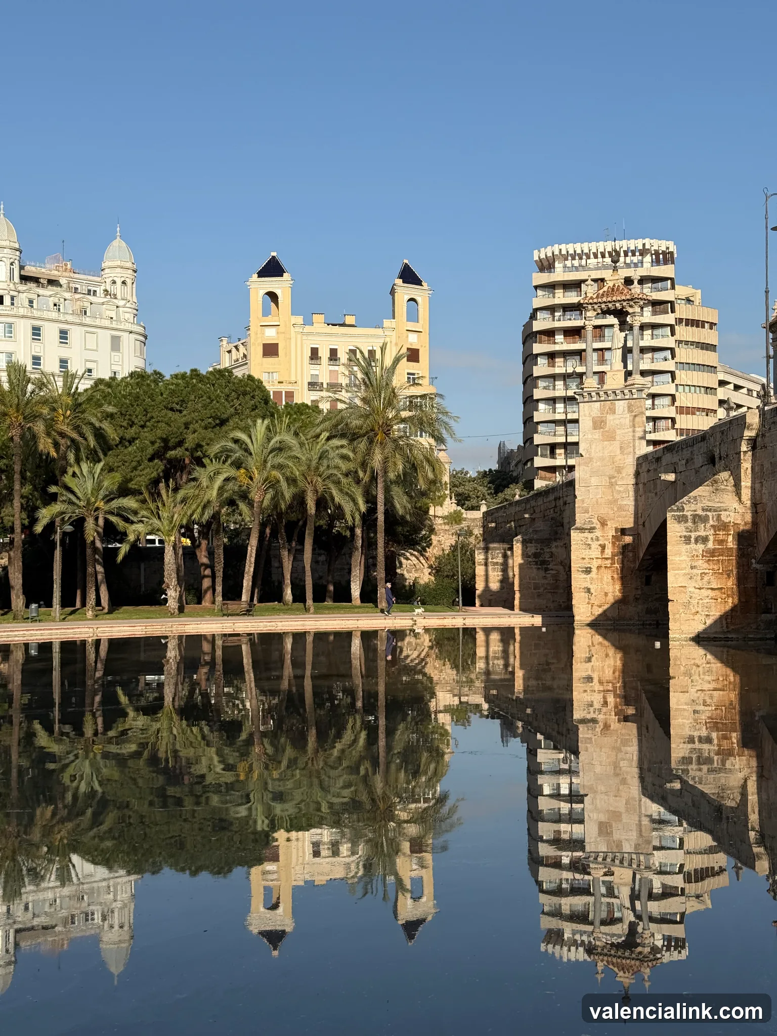 Palm trees and buildings reflected in calm water under a clear blue sky.